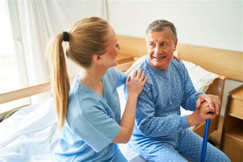 Caring Nurse Sitting With Elderly Man On Bed At Rehabilitation Center