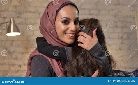 Young Beautiful Mother In Hijab Hugs Her Little Daughter Looks At Camera Sitting On Couch