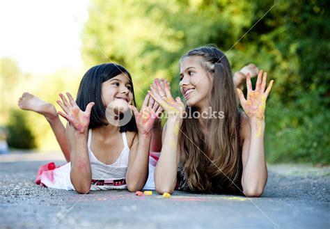 Two Sisters Laughing And Playing With Chalks On Pavement In Green Sunny