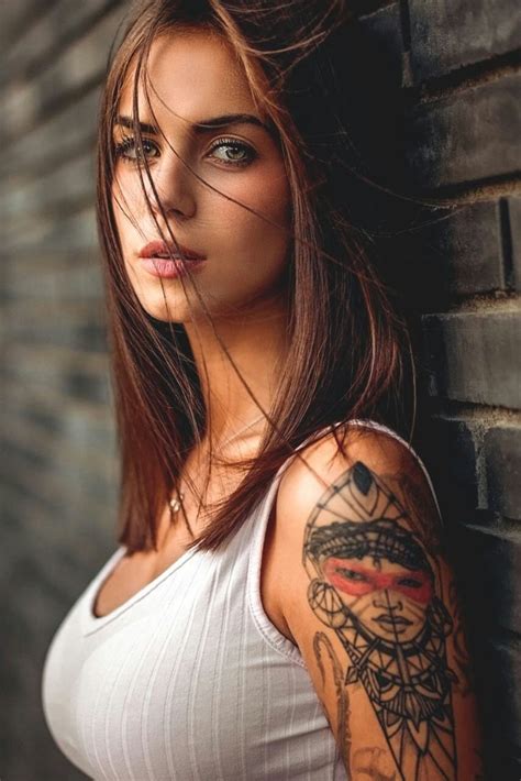 A Woman With Tattoos On Her Arm Leaning Against A Brick Wall And Looking At The Camera