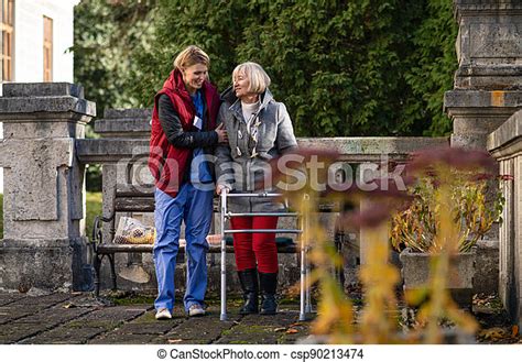 Senior Woman With Walking Frame And Caregiver Outdoors On A Walk In