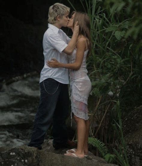 A Man And Woman Standing Next To Each Other In Front Of A River With Trees