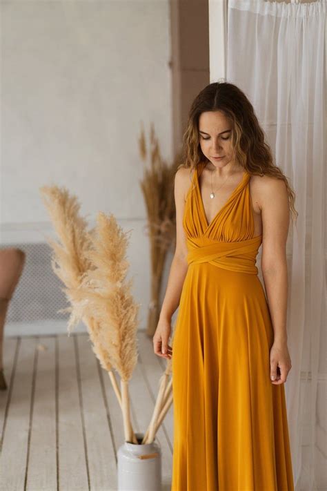 A Woman In A Long Yellow Dress Standing Next To A Tall White Vase With Dry Grass