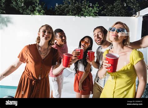 Interracial People With Plastic Cups Looking At Camera Near Swimming Pool At Night Stock Photo