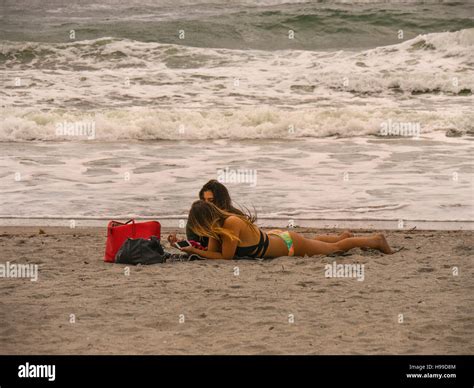 Two Tanned Girls Lying On The Beach By The Sea Looking At Their Mobile