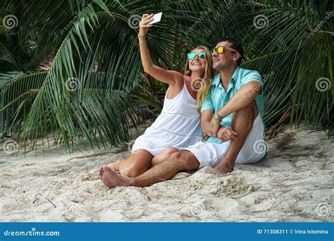 The Young Couple The Woman And The Man Sit On The Beach And Will Take The Selfies Dressed In