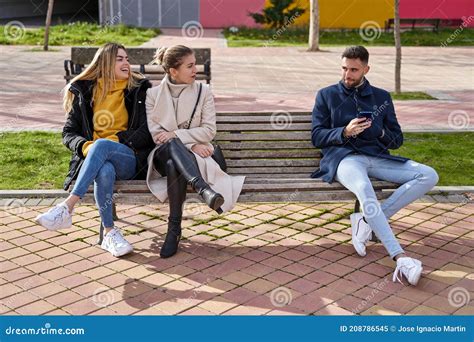 Two Blonde Girls Sitting On A Bench Next To A Guy Who Is Using His Cell Phone They Are Looking