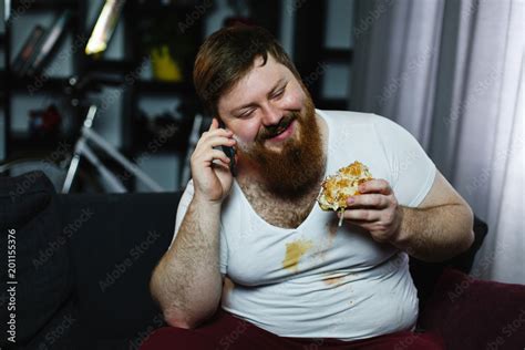 Ugly Fat Man Talks On His Smarphone While He Eats A Burger Stock Photo