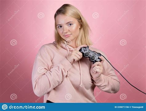 Charming Blonde Gamer A Young Smiling Woman In A Pink Hoodie Holds A