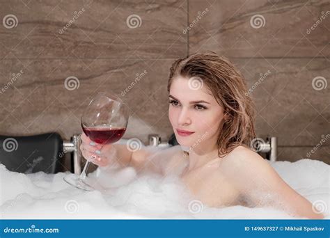 Attractive Blonde Woman Lying In Bathtub In Foam In Hotel Drinking Red Wine From A Large Glass