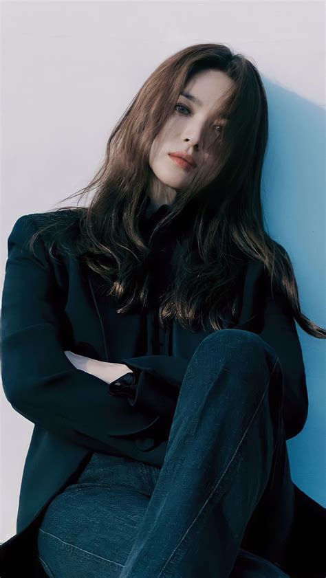 A Woman With Long Hair Sitting On The Ground Wearing Jeans And A Black Coat Is Looking At The Camera
