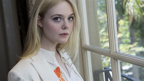 Mary Elle Fanning Is Standing With White Shirt And Coat Near Window