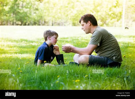 Side View Of Father And Son Sitting On Grassy Field At Park Stock Photo