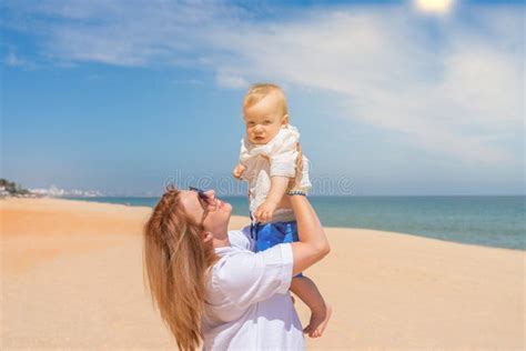 Young Mother In A White Dress On Beach Near Sea Holds Her Baby Son In