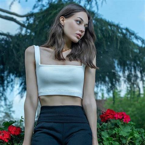 a woman in black pants and a white crop top posing for the camera with red flowers behind her