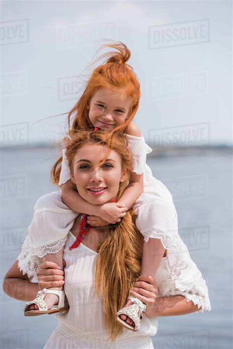 happy mother carrying adorable redhead daughter on neck and smiling at camera at seashore