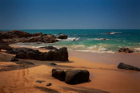 View Of Rocks On Sand Beach Wave Surf Against Azure Sea Sky Stock Photo