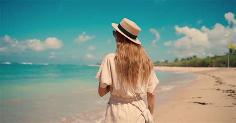 romantic blonde woman tourist in straw hat walking to ocean sandy beach in sunny day rear view