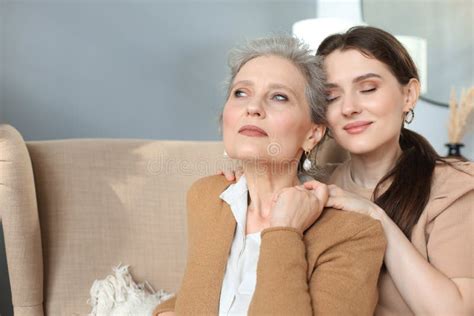 Happy Daughter Hugging Older Mother Standing Behind Chair In Living