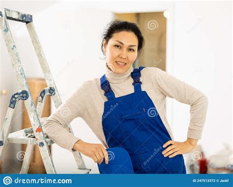 Portrait Of Positive Builder Woman In Blue Overalls Next To Stepladder