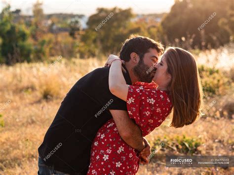 Man Kissing Smiling Pregnant Wife On Background Of Picturesque Green Nature In Sunny Day