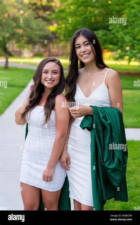 Two College Girls And Best Friends Walk On A Sidewalk While Holding Their Caps And Gowns Before