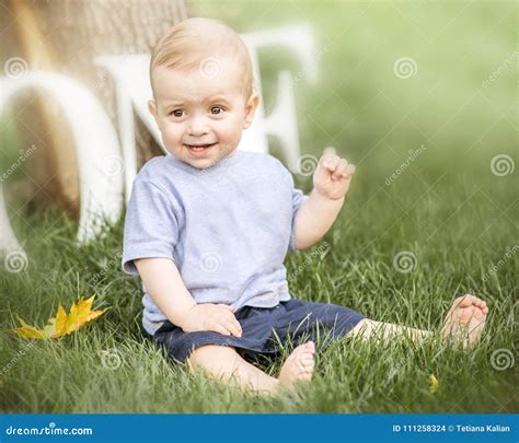 A Portrait Of A Happy Cute Baby Boy Sitting On Green Grass Outdoor At