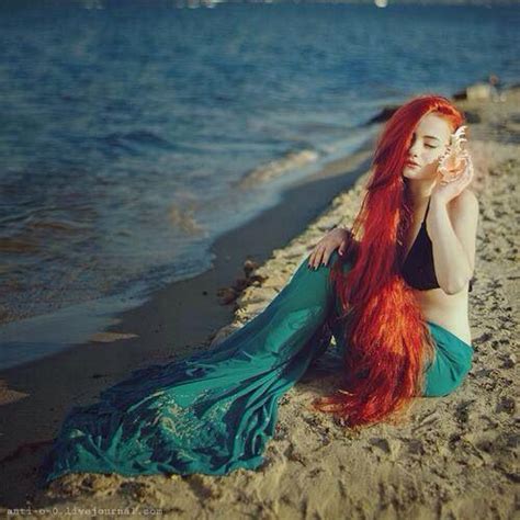 Mermaid With Long Red Hair Green Tail Sitting On The Sand Holding A Seashell To Her Ear
