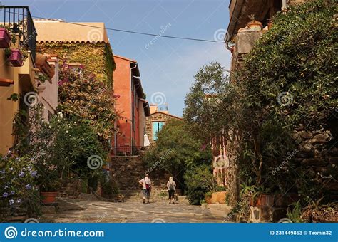 Two People Walking On Street Bordered By Greenery And Colourful Houses