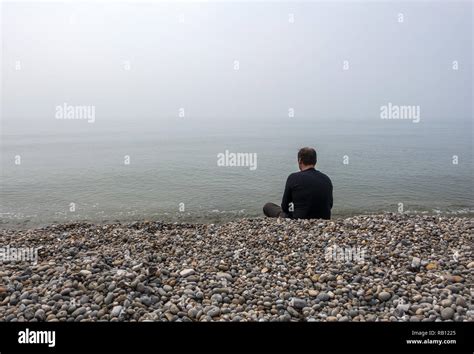 Man Sitting Alone On Pebble Beach Looking Out Into The Sea Covered By