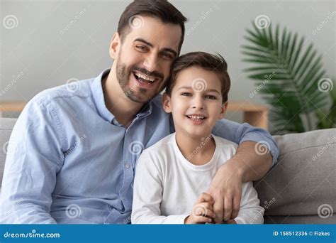 Happy Dad Embracing Son Looking At Camera Sit On Sofa Stock Photo