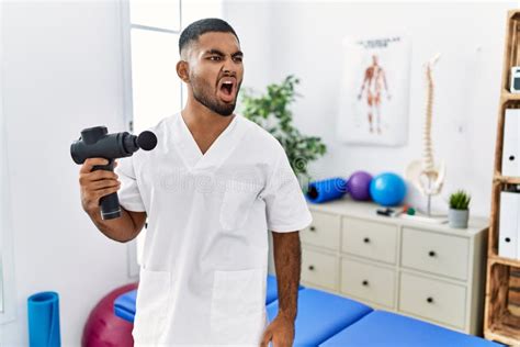 Young Indian Physiotherapist Holding Therapy Massage Gun At Wellness Center Angry And Mad