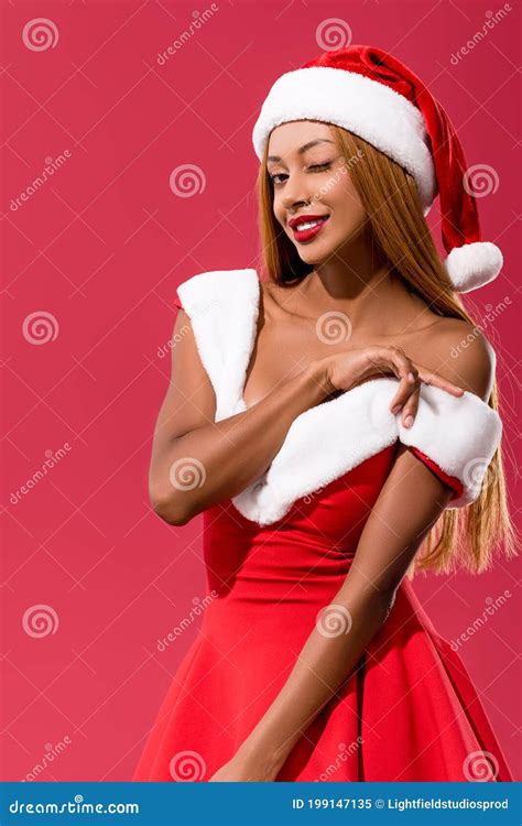 African American Girl In Santa Hat Baring Shoulder And Winking At Camera Isolated On Red Stock