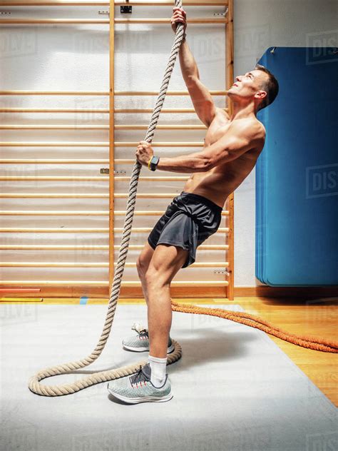 Side View Of Shirtless Muscular Sportsman Climbing Up Rope While