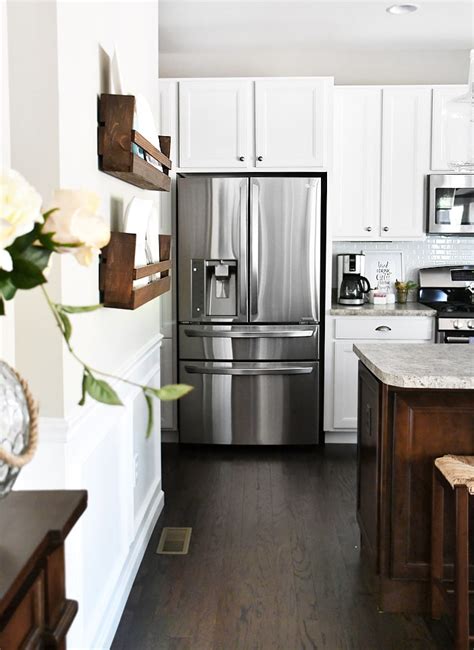 In a corner beside a pantry door, a white and gray kitchen boasts a light blue china cabinet finished with antiqued mirrored doors, as darlana lanterns hang over a white kitchen island finished with a dark gray quartz countertop seating brown wicker stools on a dark brown wide plank wood floor. White Cabinets, Dark Kitchen Island - How to Nest for Less™