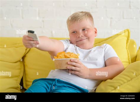Obese Boy Eating Chips Resting On A Sofa And Turning Tv On Stock Photo