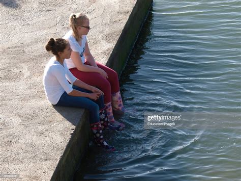 Two Teenage Girls Sat On The Edge Of Sea Pool Dangling Feet In The