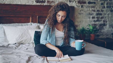 Pretty Girl Reading Book Sitting On Bed At Home And Holding Cup With