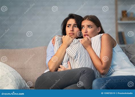Two Scared Girls Hugging Pillows Sitting On Couch At Home Stock Image