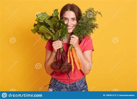 Smiling Brunette Female Hiding Behind Carrots And Beets In Her Hands Posing In Casual Attire