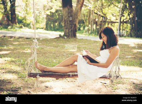Beautiful Asian Woman In A White Dress Sitting On A Wooden Swing Reading A Book In The Park