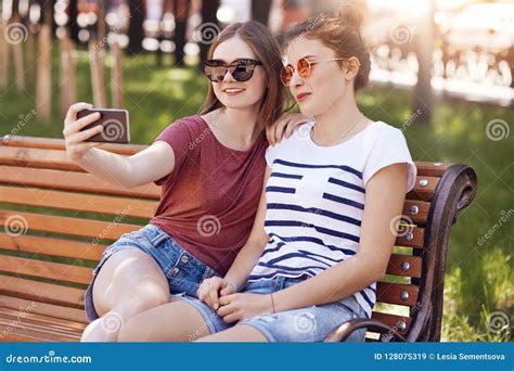 Joyful Two Girls Make Selfie Portrait With Modern Cell Phone Sit Closely To Each Other On Bench