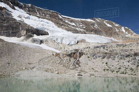 Glacier On Mountain Side Streaming Down Into A Reflective Mountain Pond