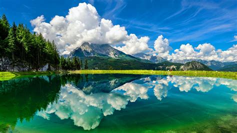 Landscape View Of Rock Mountains Under Beautiful White Clouds Reflection On River Green Grass