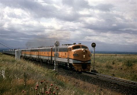 An Orange And Silver Train Traveling Down Tracks Next To Tall Grass On