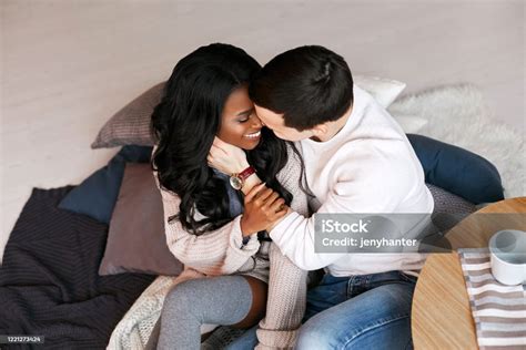 Young Interracial Couple Sitting On The Floor Hugging And Kissing The Black Girl Holds The Hand