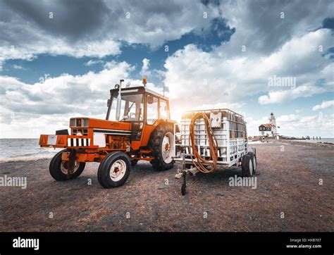 Red Tractor With Trailer Against Lighthouse And Blue Overcast Sky In
