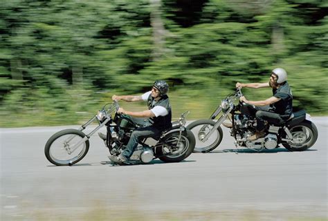 two men riding motorcycles on a road with trees in the background