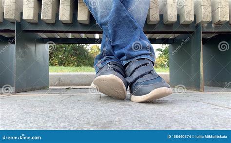 Legs Feet Of A Bored Teen Boy Sitting On A Bench In Urban City Park And