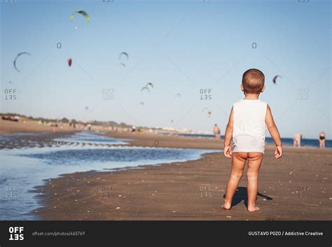 Back View Of Baby With Naked Bottom Standing On Beach Watching Flying